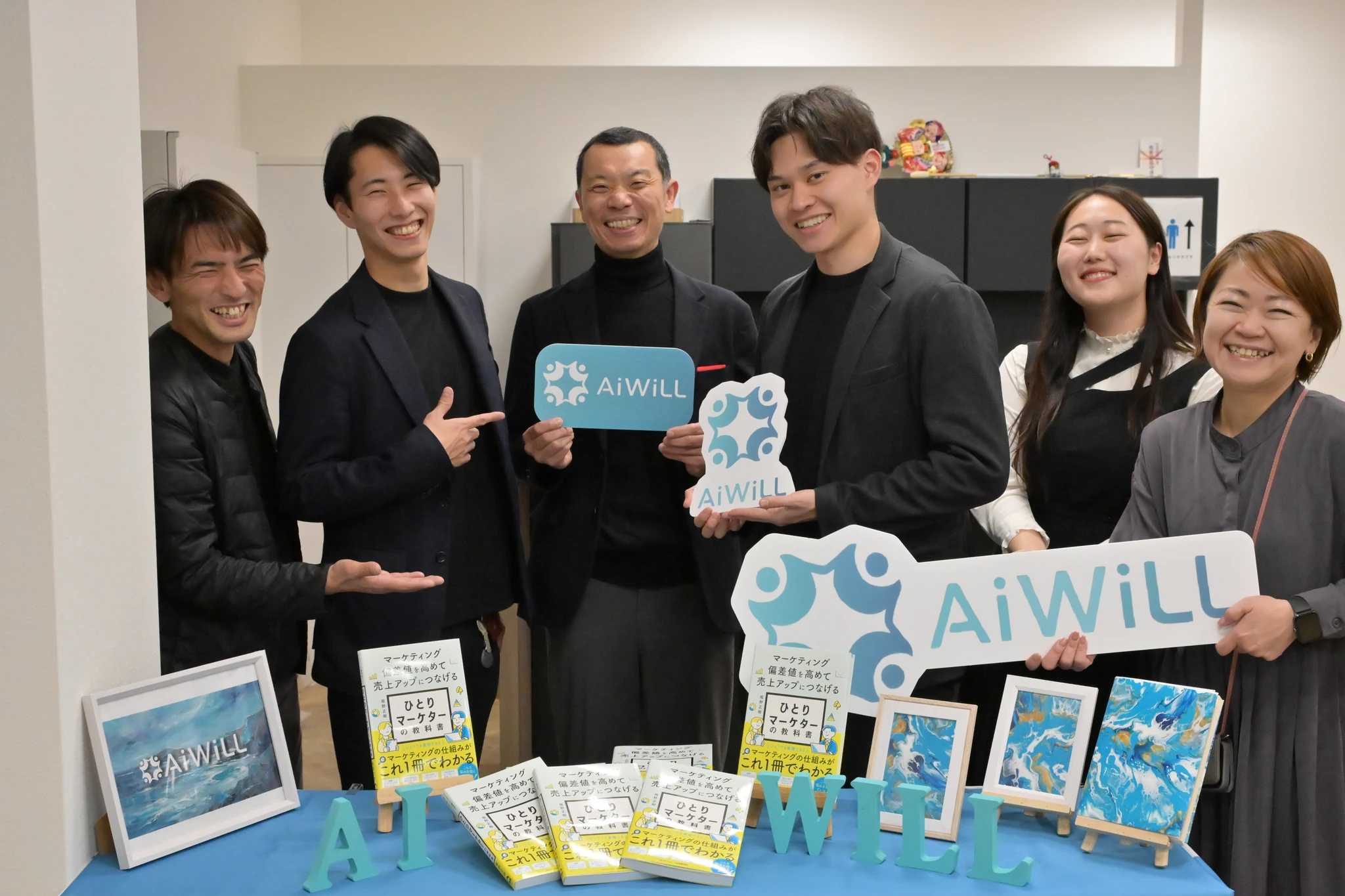AiWiLLのボードを持ち展示机を囲むメンバーの集合写真